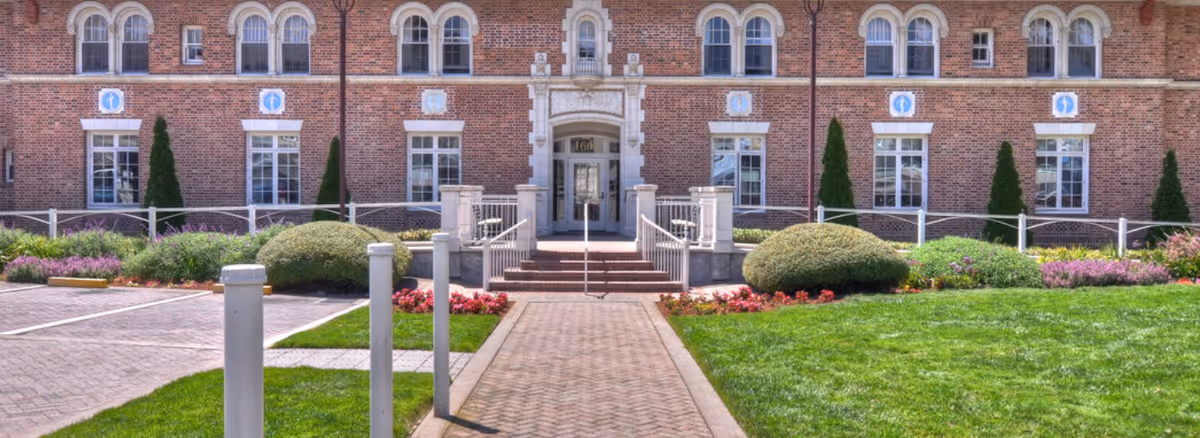 Front entrance of a brick building with a central walkway, steps, railings, and landscaped lawns and shrubs.