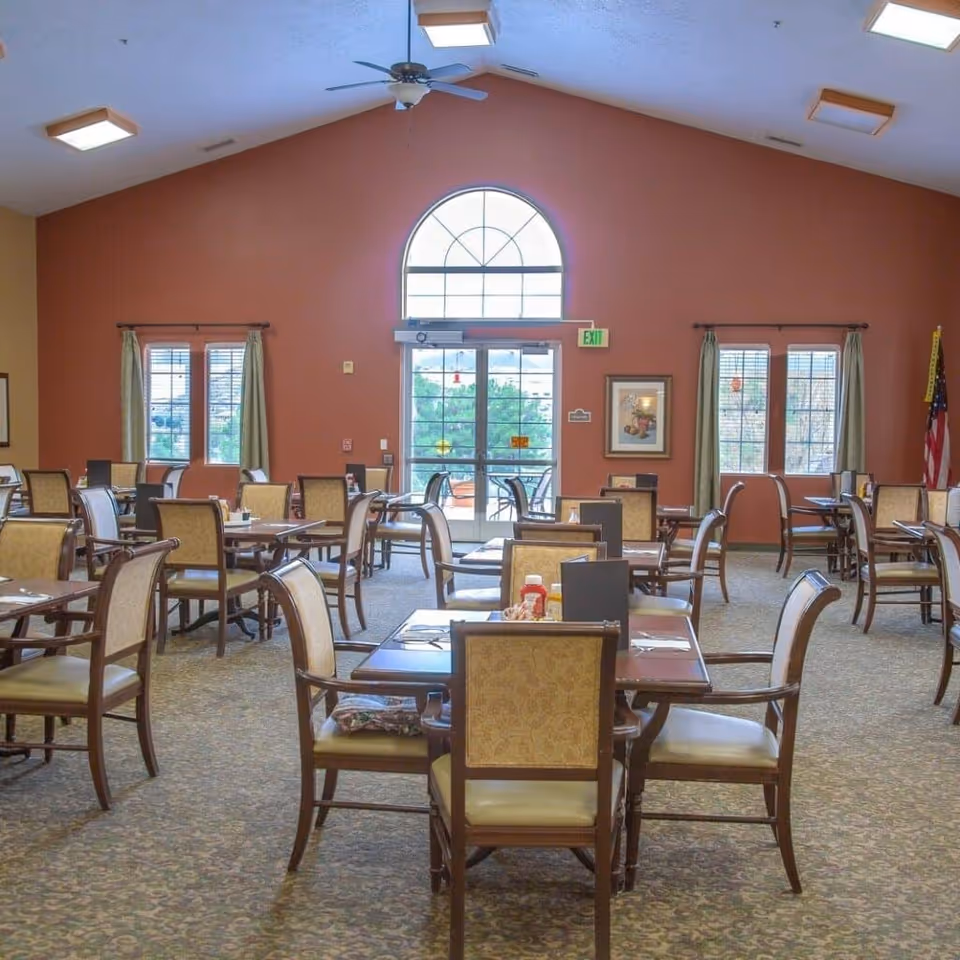 A spacious dining room with multiple wooden tables and cushioned chairs arranged neatly. The room has a high ceiling with a ceiling fan and several square light fixtures. There are large windows with curtains and a glass door with an arched window above it, allowing natural light to enter. The walls are painted in warm tones, and there is an American flag in the corner.