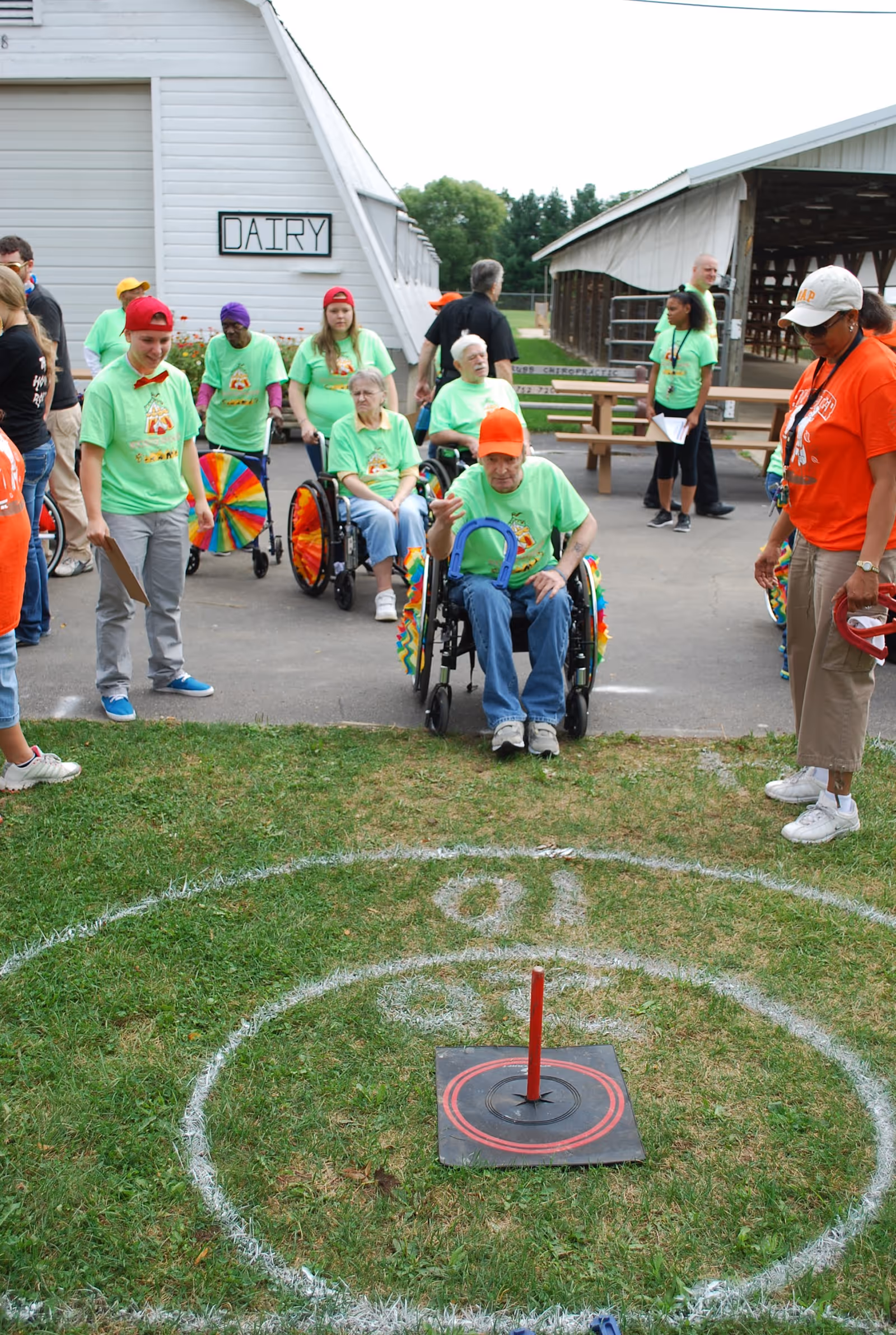 A group of elderly people, some in wheelchairs, and caregivers participate in an outdoor ring toss game near a building labeled 'DAIRY'. The participants and caregivers wear bright green and orange shirts, and the setting appears to be a farm or rural area with barns in the background.