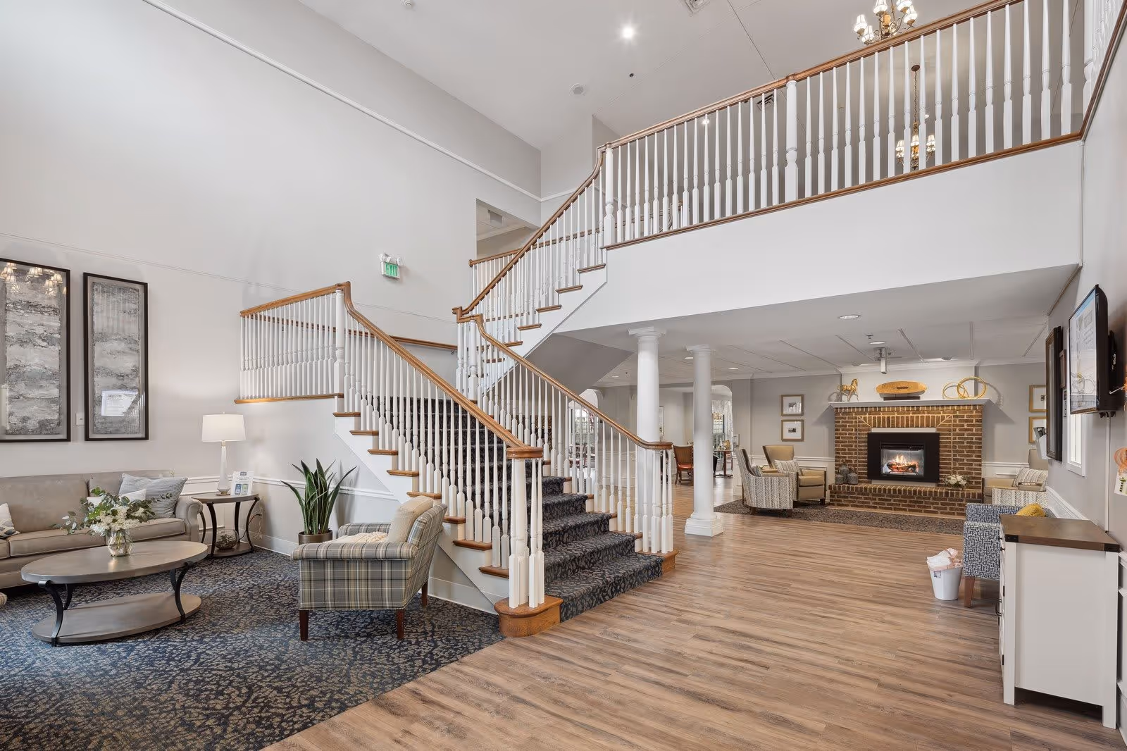 A spacious and well-lit living area in a senior living facility featuring a staircase with white railings and wooden handrails. The room includes comfortable seating with a sofa, armchairs, a coffee table, and a side table with a lamp. There is a brick fireplace with decorative items on the mantel, wooden flooring, and framed artwork on the walls.
