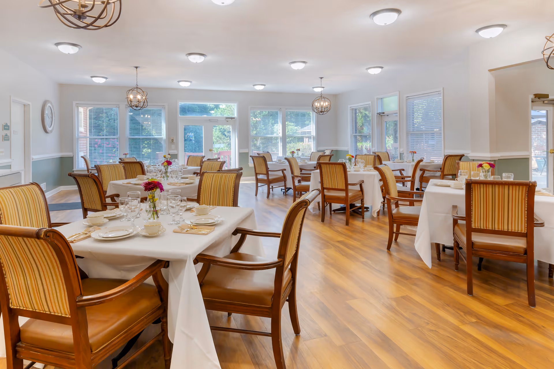 A bright and spacious dining room with multiple tables covered in white tablecloths, each set with plates, cups, glasses, and silverware. The room features wooden chairs with striped upholstery, large windows allowing natural light, and decorative hanging light fixtures. The floor is wooden, and there are doors leading outside.