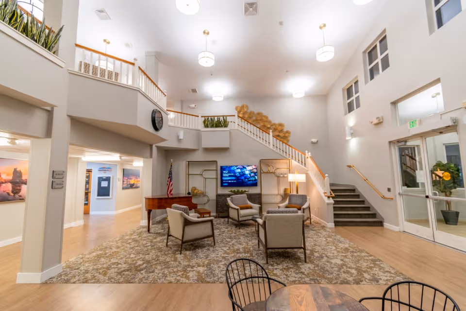A spacious and well-lit common area in a senior living facility featuring a seating arrangement with four armchairs around a small table on a patterned rug. There is a flat-screen TV mounted on the wall, a piano, decorative shelving, and a staircase leading to an upper level. The room has high ceilings with hanging light fixtures, large windows, and a glass door entrance.