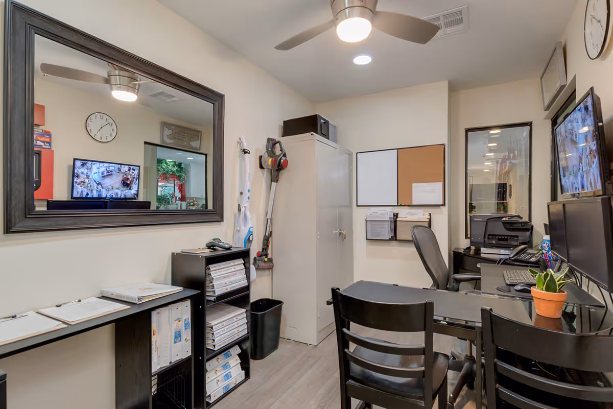 Small office space with a desk, two chairs, a computer monitor, a printer, and a potted plant. There is a large mirror on the wall reflecting a clock and a TV screen showing multiple security camera feeds. Shelves with binders and clipboards are also visible, along with a white cabinet and cleaning equipment mounted on the wall.