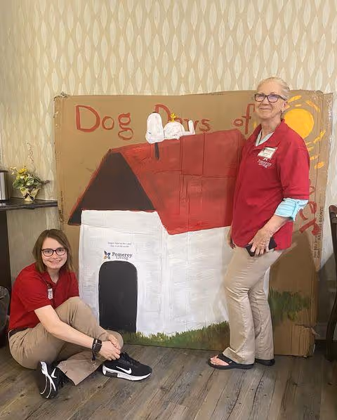Two women wearing red shirts with Pomeroy Living logos pose indoors next to a large painted cardboard cutout of Snoopy's doghouse. One woman is sitting on the floor smiling, and the other is standing. The background includes a patterned wallpaper and a small table with a plant.