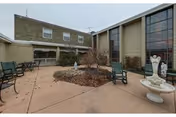 Outdoor courtyard area at a senior living facility with a circular garden bed in the center, surrounded by benches and chairs. The courtyard is enclosed by two-story buildings with large windows and beige siding. A decorative white fountain with angel statues is visible on the right side.