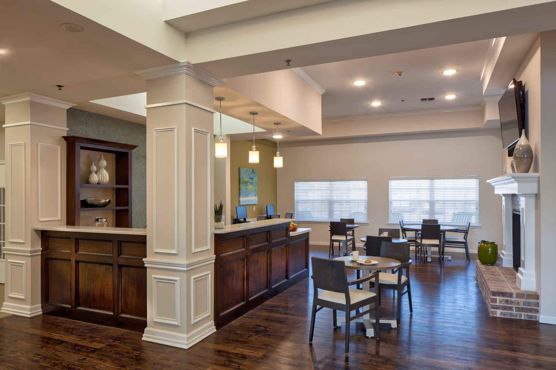 Interior view of a common area in an assisted living facility featuring a reception desk with pendant lights, several tables and chairs, a fireplace with a TV mounted above it, and large windows allowing natural light to fill the space.
