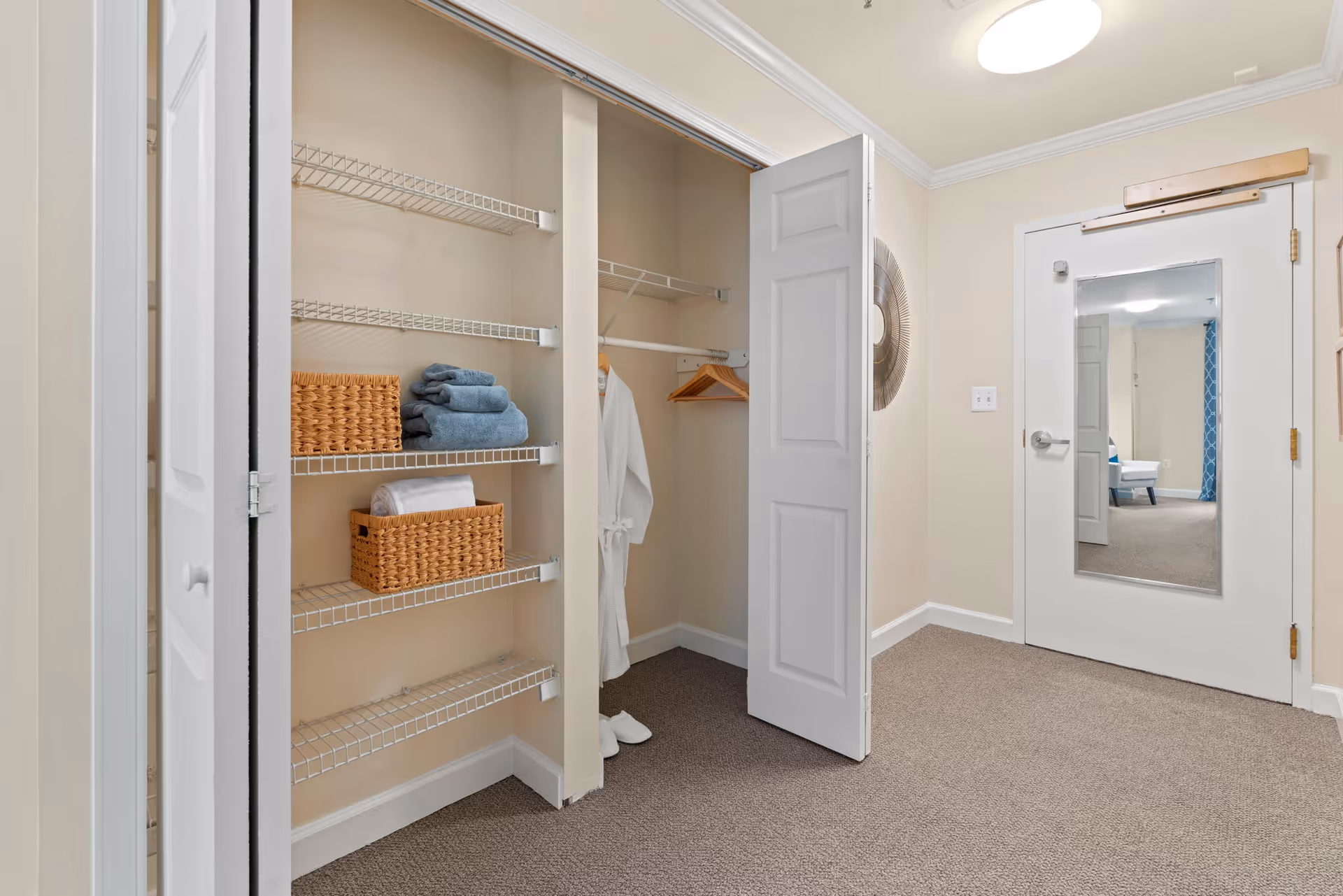 Interior view of a room with an open closet featuring wire shelves holding folded towels and baskets, a hanging white robe and slippers on the floor. A door with a full-length mirror reflects part of another room with a chair and blue curtains. The walls are beige and the floor is carpeted.