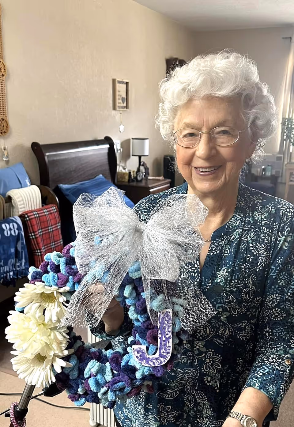 An elderly woman with white curly hair and glasses is smiling while holding a decorative wreath adorned with white flowers, a large silver bow, and a purple letter J. She is standing in a bedroom with a dark wooden bed, blue and red blankets, a nightstand with a lamp, and other bedroom furnishings visible in the background.