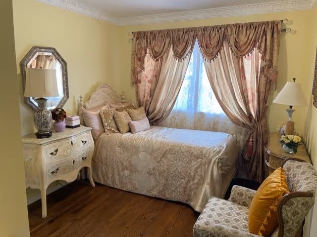 A cozy bedroom with a single bed covered in a beige patterned bedspread and multiple decorative pillows. The bed is positioned against a window with ornate, heavy drapes in shades of gold and pink. To the left of the bed is a vintage-style white dresser with a lamp, a small plant, and a box on top. To the right, there is a small round table with a lamp and a vase of flowers, and in front of it, an upholstered armchair with a mustard yellow pillow. The room has wooden flooring and pale yellow walls.