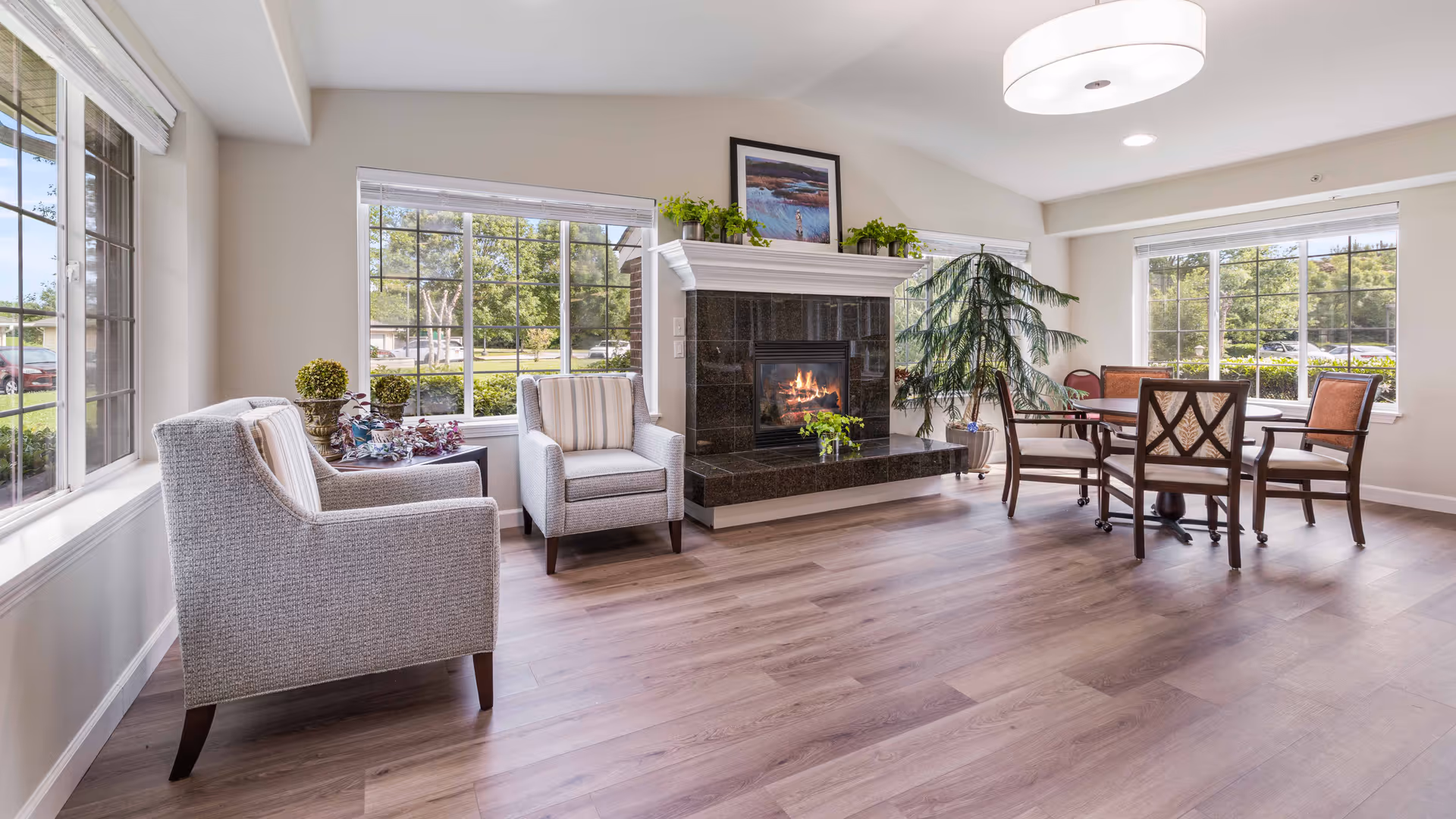 A bright and spacious living room area with large windows showing outdoor greenery. The room features two upholstered armchairs, a black marble fireplace with a fire burning, a framed picture and plants on the mantel, a round wooden table with four chairs, and a large potted plant near the windows. The floor is light wood, and a modern ceiling light fixture is visible.
