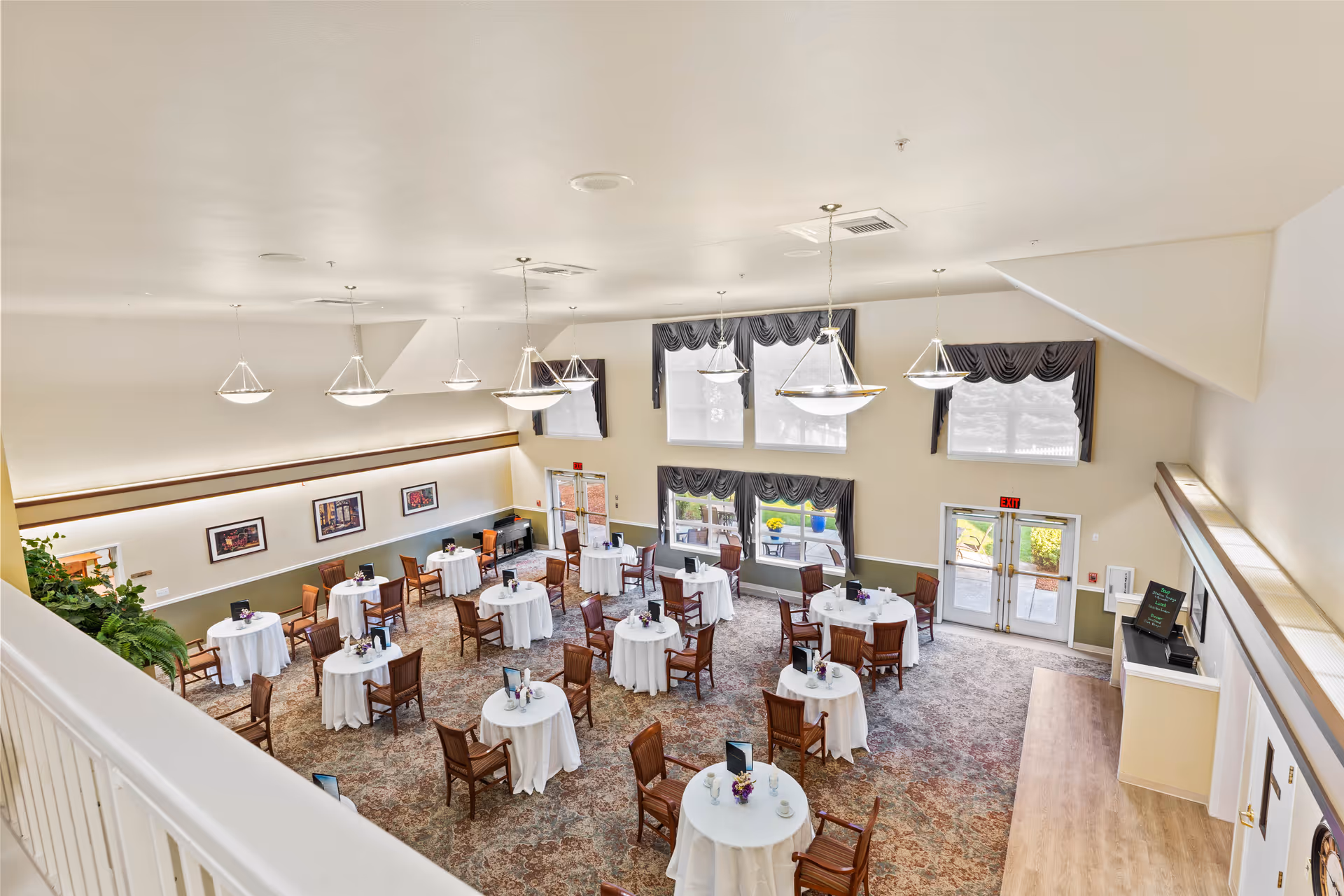A spacious dining room with multiple round tables covered with white tablecloths, each surrounded by wooden chairs. The room has large windows with dark valances, hanging pendant lights, framed artwork on the walls, and a carpeted floor. There is a door leading outside and a small counter area on the right side.