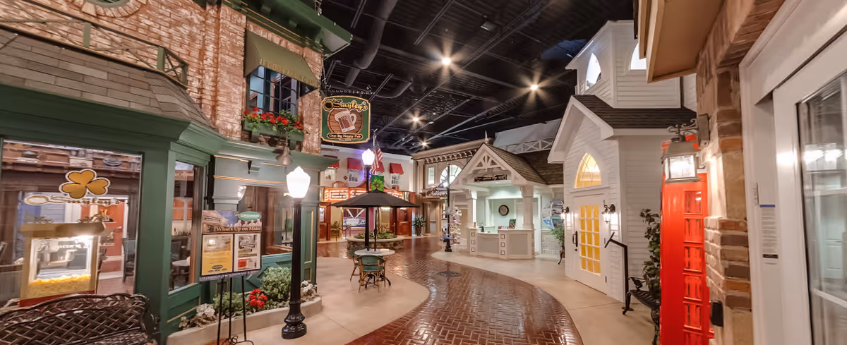 Indoor street-like setting in a senior living facility with storefront facades, including a green building with a shamrock sign, a white building with a reception desk, a red telephone booth, and a central seating area with a table and umbrella under street lamps.