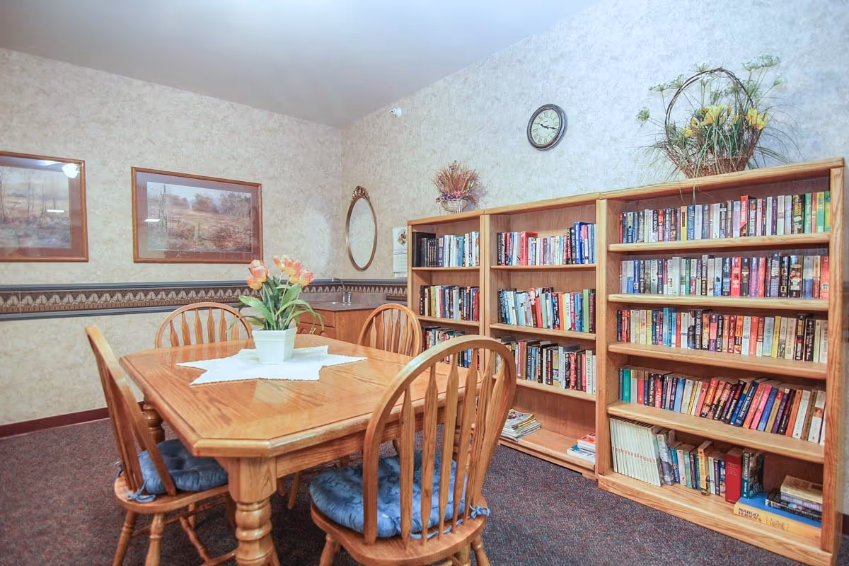 Community room with a wooden dining table and chairs, a potted flower centerpiece, and bookshelves filled with books.