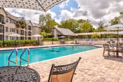 Outdoor swimming pool area at Veranda Club with lounge chairs, tables with umbrellas, and residential buildings in the background under a partly cloudy sky.