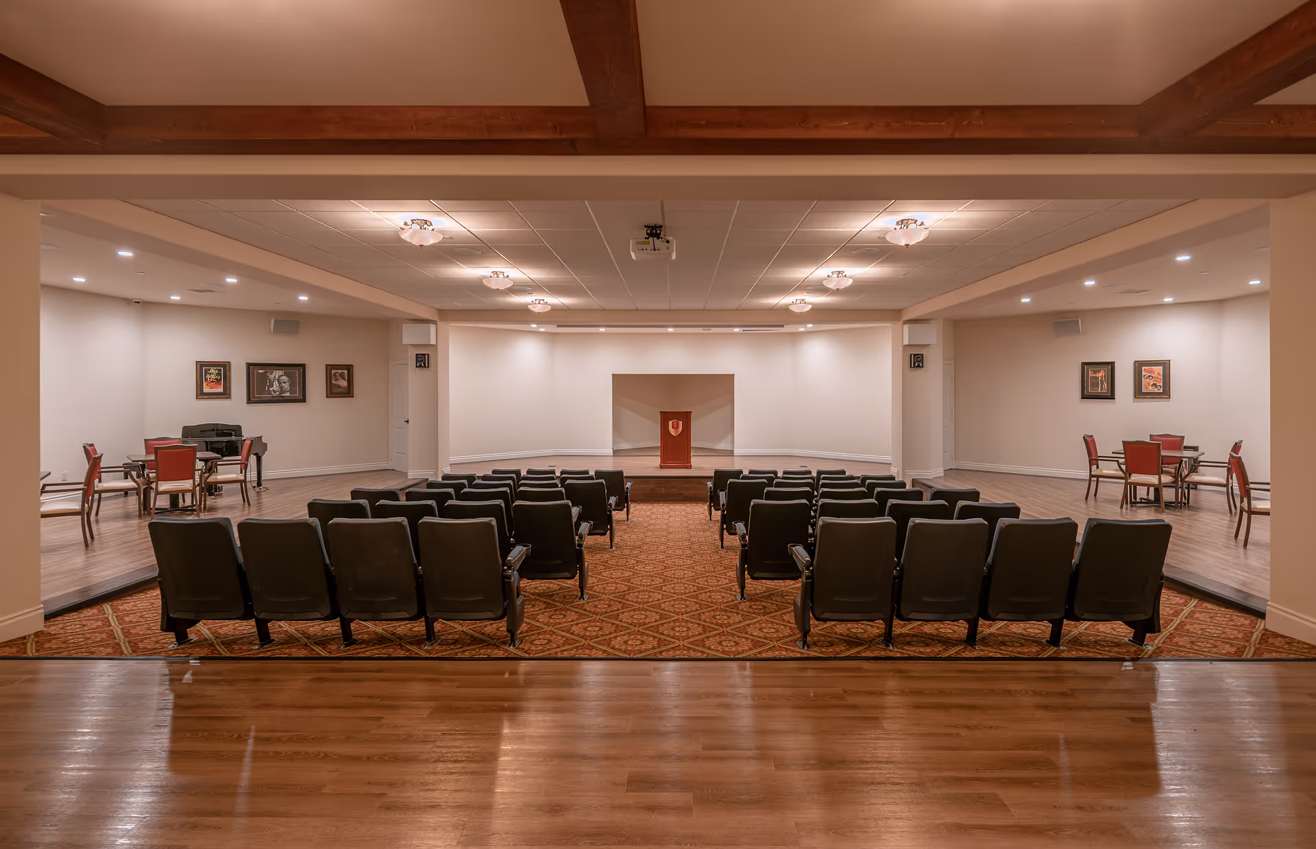 A spacious auditorium or meeting room with rows of black chairs facing a small stage with a wooden podium. The room has wooden flooring, a patterned carpet in the seating area, and a ceiling with recessed lighting and wooden beams. On either side of the room, there are small seating areas with tables and red chairs, along with framed pictures on the walls.