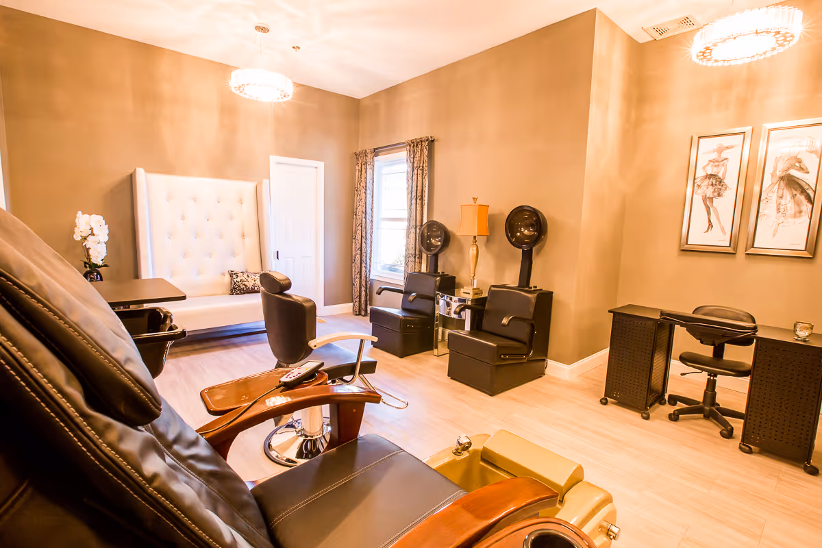 Well-lit salon room with pedicure and salon chairs, hair dryers, a manicure station, and a tufted bench.