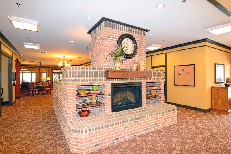 Interior common area featuring a large central brick fireplace topped with a clock, with seating and a dining area visible in the background.