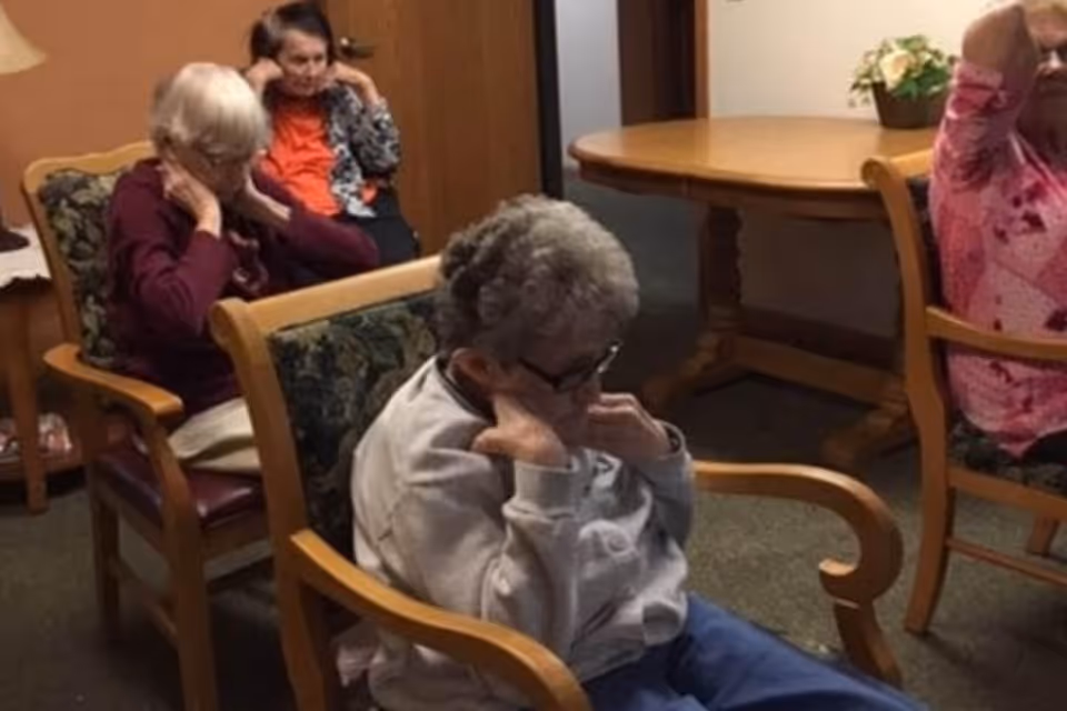 A group of elderly individuals sitting in wooden chairs with floral cushions in a room with a round wooden table and a potted plant in the background. They appear to be engaged in a quiet activity, with some covering their ears.