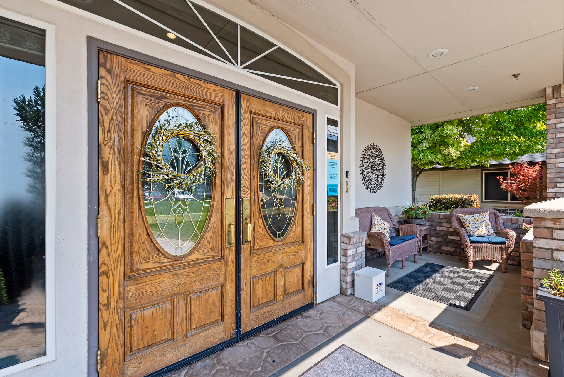 Entrance to a facility with double wooden doors featuring oval glass panels decorated with wreaths. To the right of the doors is a covered porch area with two wicker chairs with cushions, a small table, a decorative wall piece, and some plants. Trees and bushes are visible in the background.
