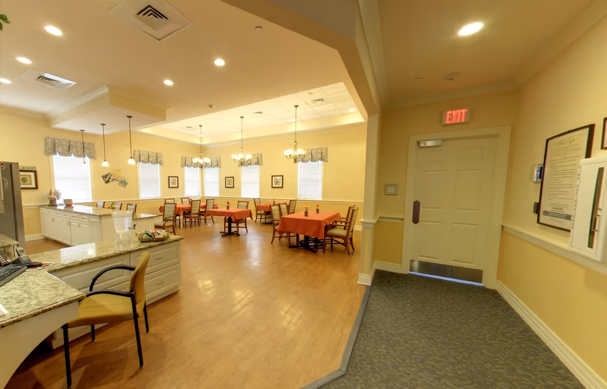 Bright communal dining room with multiple tables covered in orange tablecloths, chairs, a reception desk in the foreground, and an exit door to the right.
