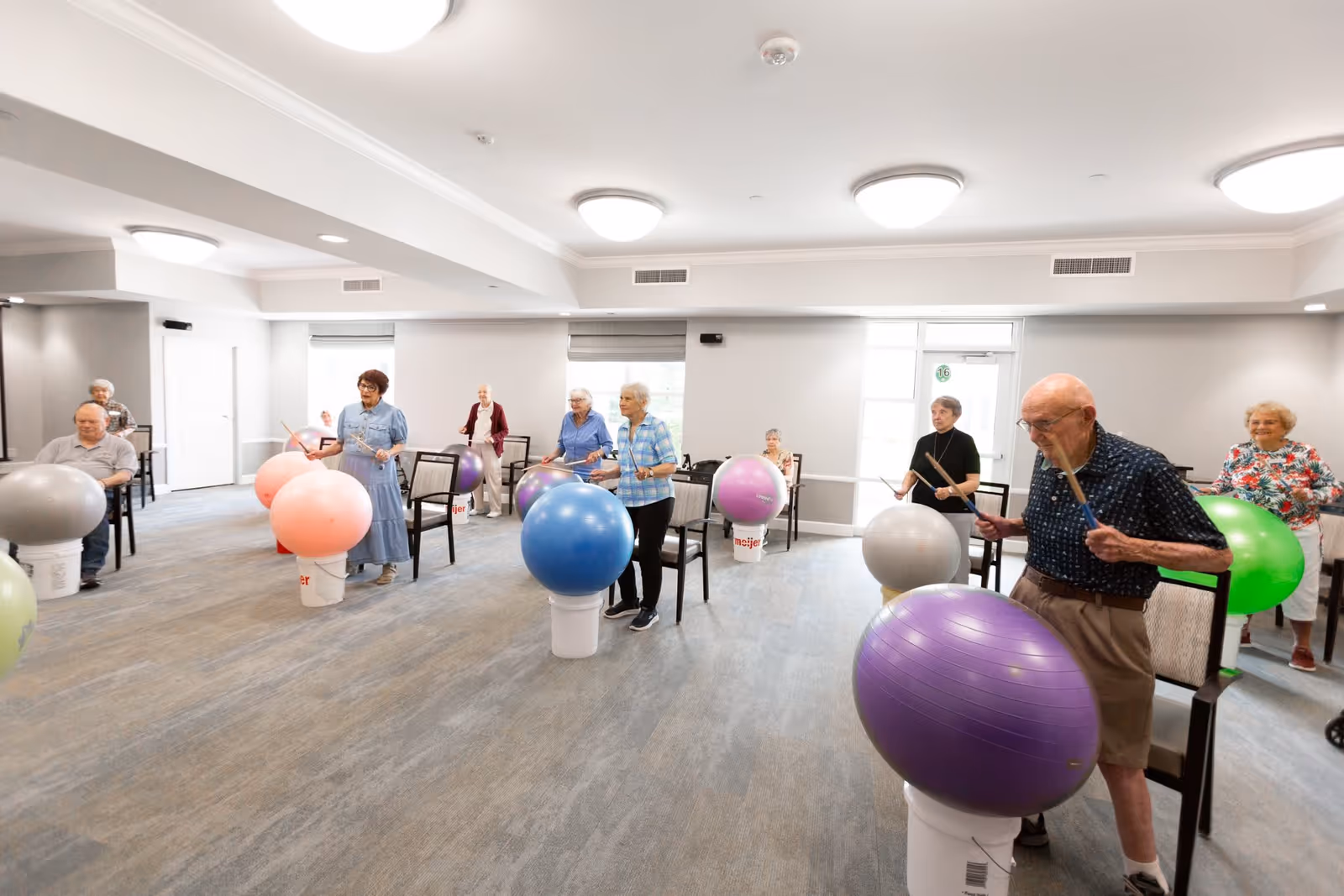 A group of elderly people participating in a fitness or recreational activity indoors, using large colorful exercise balls placed on white buckets and drumsticks to hit the balls. The room is spacious with chairs arranged around the participants and has bright lighting from ceiling fixtures and windows.