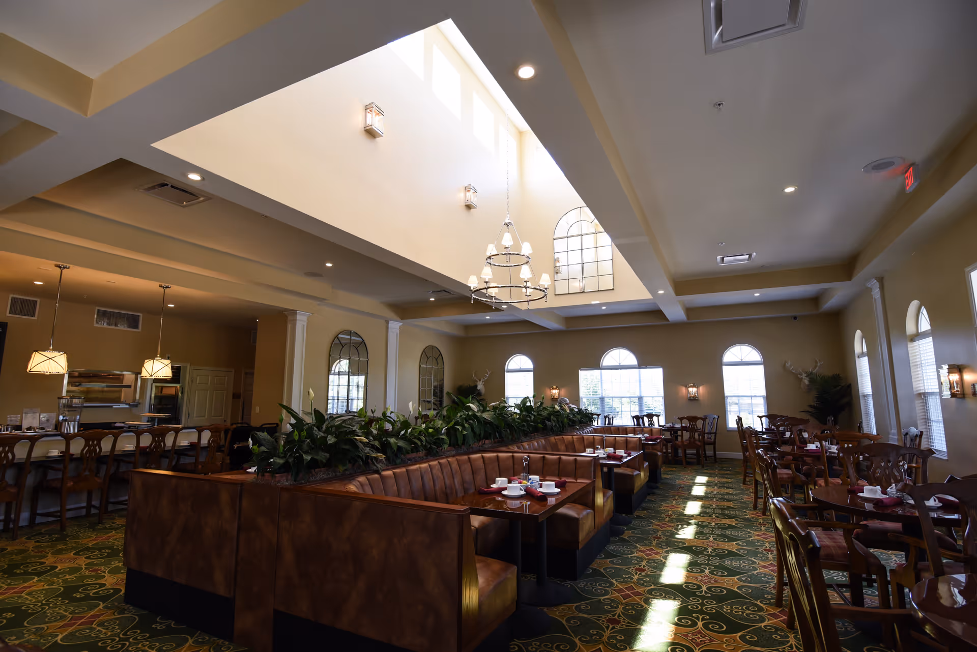 A spacious dining room with a high ceiling and large skylight. The room features a long brown leather booth seating area with tables set with cups and saucers. There are additional wooden chairs and tables along the windows, which have arched tops. The room is decorated with plants, wall sconces, and a chandelier hanging from the ceiling.