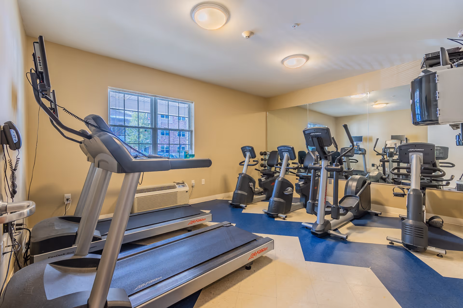 A small fitness room with two treadmills, two stationary bikes, and an elliptical machine. The room has beige walls, a large mirror on one side, a window with a view of a building outside, and blue and beige patterned flooring.