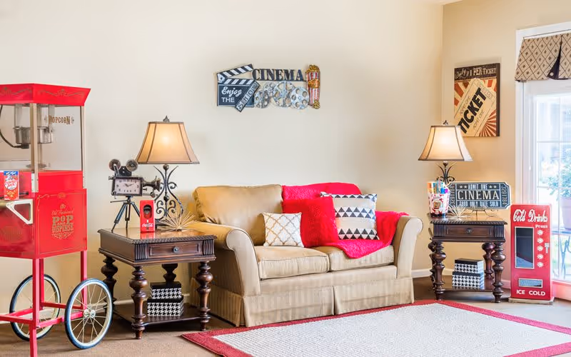 A cozy living room area with a beige loveseat adorned with red and patterned pillows and a red throw blanket. On either side of the loveseat are dark wooden side tables with decorative lamps. To the left, there is a vintage-style red popcorn machine, and to the right, a red cold drinks vending machine. The walls have movie-themed decorations including a 'Cinema' sign and vintage ticket posters. A large window with a patterned valance lets in natural light, and a red-bordered beige rug covers the floor.