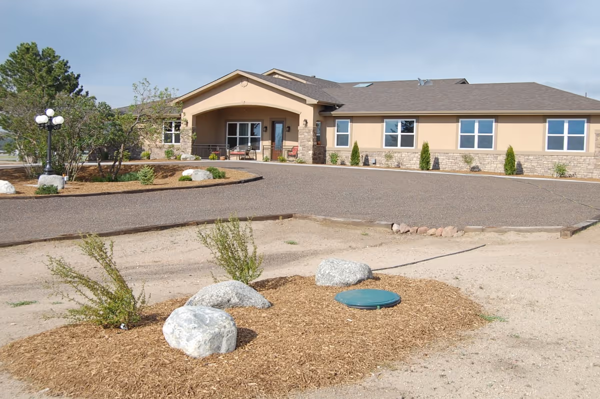 Exterior view of a single-story assisted living facility building with beige walls and a brown roof. The building has several windows and a covered entrance with a small seating area. The surrounding area includes a paved driveway, some landscaping with bushes, rocks, and a lamp post, and a mulched area with small plants and large rocks in the foreground.