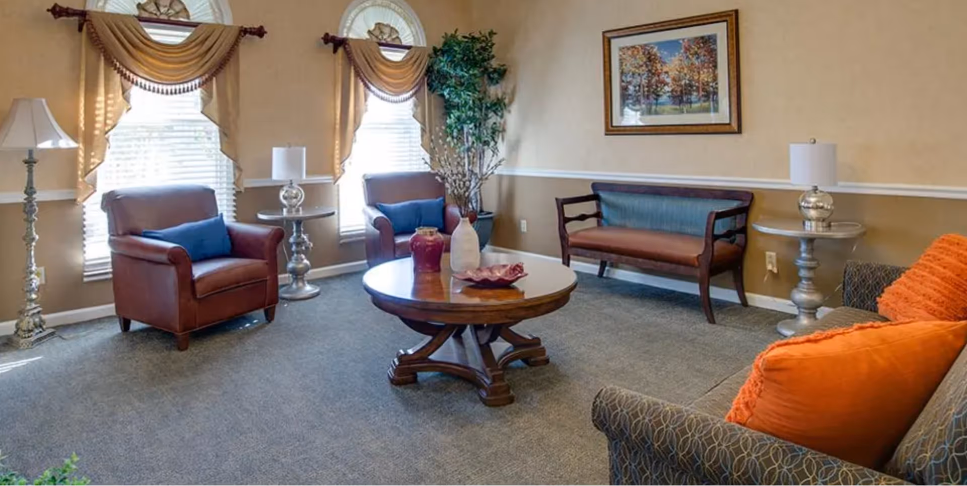 A cozy senior living room with two brown leather armchairs with blue pillows, a wooden bench with blue upholstery, a patterned sofa with orange pillows, a round wooden coffee table with decorative vases, two silver side tables with lamps, a tall floor lamp, and two windows with beige curtains. A framed picture of trees hangs on the beige wall.
