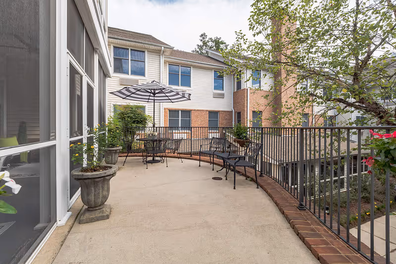 Outdoor patio area at a senior living facility with metal chairs, a table with a striped umbrella, potted plants, and a railing overlooking a garden area. The building exterior features white siding and brick accents with multiple windows.