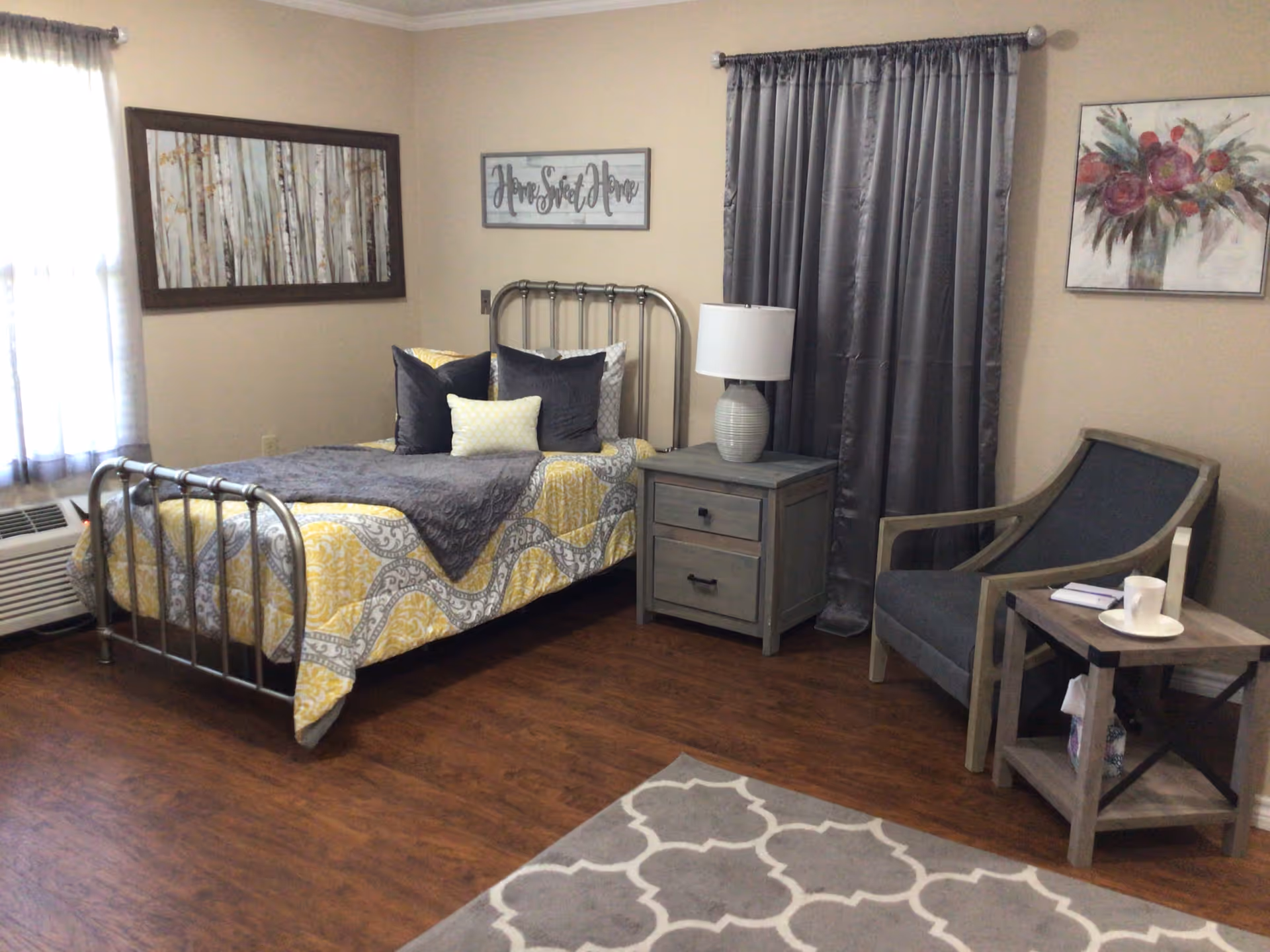 Cozy bedroom with metal bed frame, nightstand, lamp, chair, curtains, and wall art.