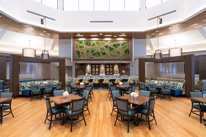 A spacious dining room with wooden floors, tables set for dining, and a green wall feature. The area is well-lit with large windows and modern decor, including floral accents on seating.