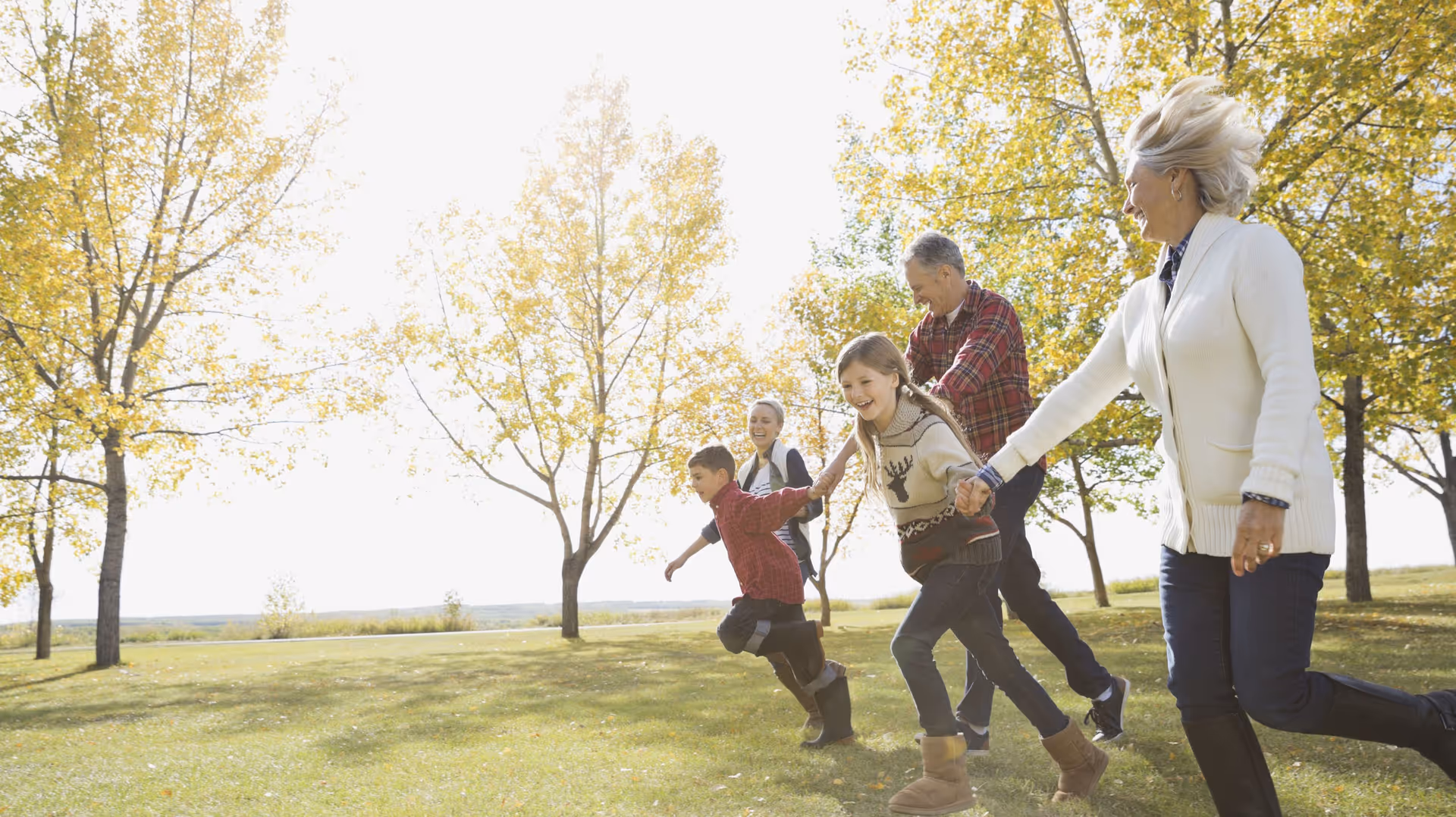 Two elderly adults and three children holding hands and running together outdoors in a grassy area with trees showing autumn foliage on a sunny day.