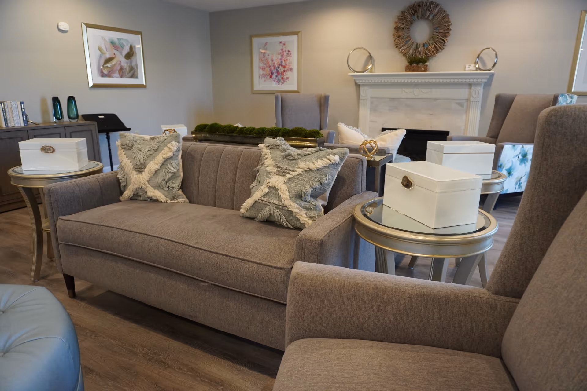 A cozy living room area with a gray sofa adorned with two decorative pillows, two matching armchairs, and round side tables with white decorative boxes on top. The room features a white fireplace with a wreath above it, framed artwork on the walls, and a wooden floor.