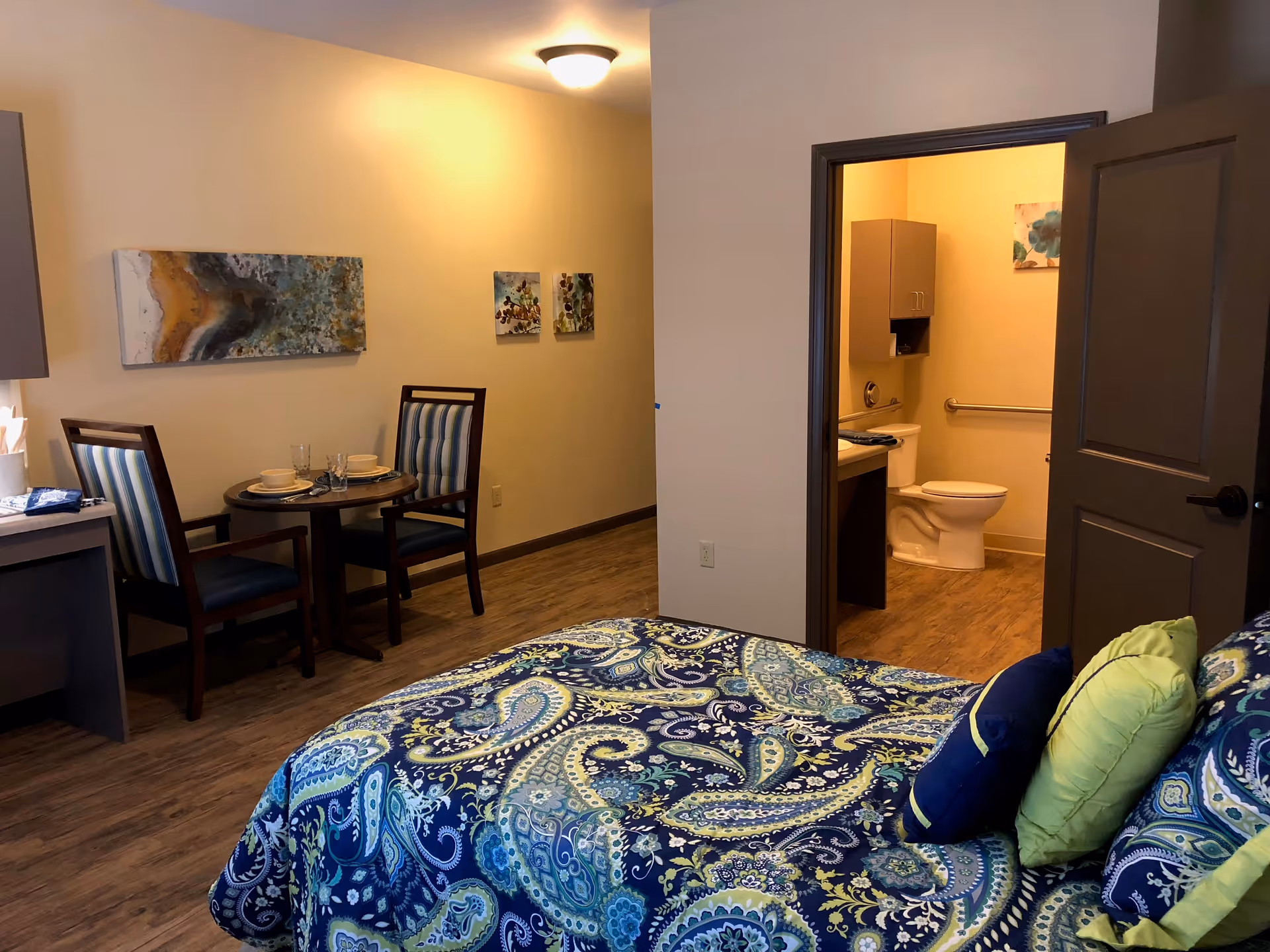 Interior view of a senior living facility room featuring a bed with blue and green paisley bedding, a small dining table with two chairs, and a bathroom with an open door showing a toilet and sink. The room has wooden flooring and light-colored walls with decorative paintings.