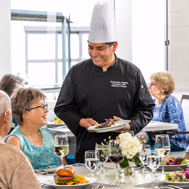 A chef wearing a black uniform and white chef hat serves a plated meal to a smiling elderly woman seated at a dining table. Other elderly people are seated around the table with various dishes and drinks, including a burger, fries, and wine glasses. The setting appears to be a bright dining room with large windows.