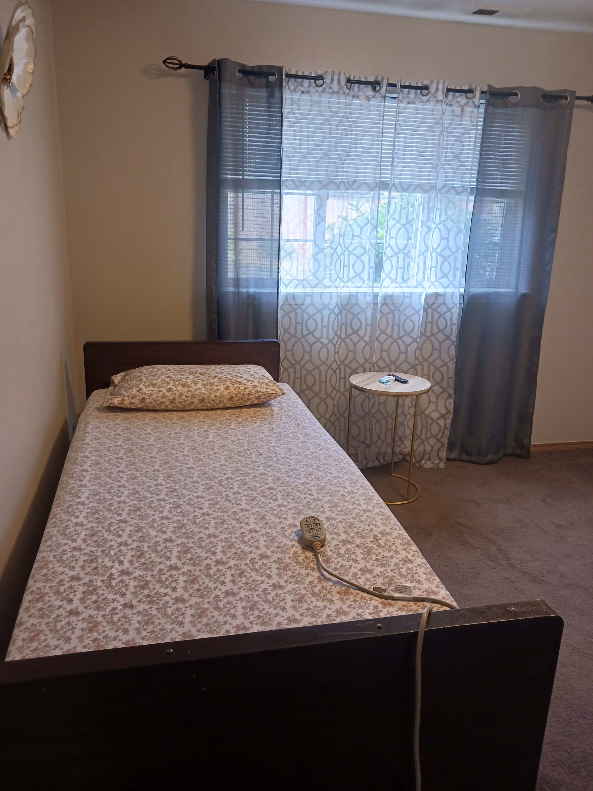 A small bedroom with a single bed covered in floral-patterned sheets and a matching pillow. A remote control is placed on the bed. Next to the bed is a small round side table with a remote and some small items on it. The window behind the table has sheer patterned curtains and darker outer curtains. The room has beige walls and carpeted floor.