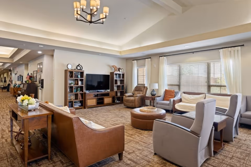 A spacious and well-lit living room area in a senior living facility featuring a brown leather sofa, two gray armchairs, a recliner, a round ottoman, and a wooden entertainment center with a flat-screen TV. Large windows with white curtains allow natural light to fill the room, and a chandelier hangs from the ceiling. The room has a warm and inviting atmosphere with carpeted flooring and neutral-colored walls.