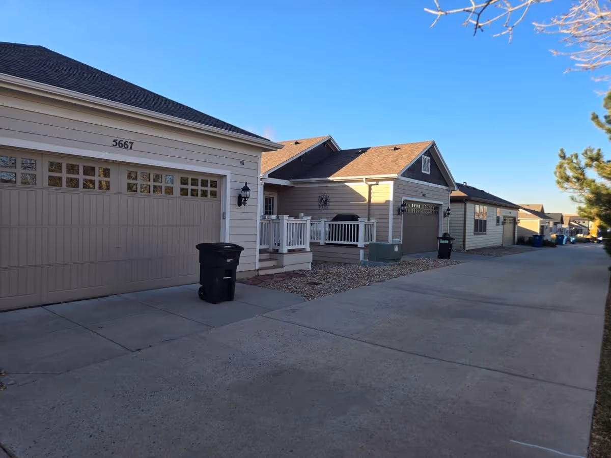 Alley and driveway lined with single-story beige garage-front homes, small porches, and trash bins under a clear blue sky.