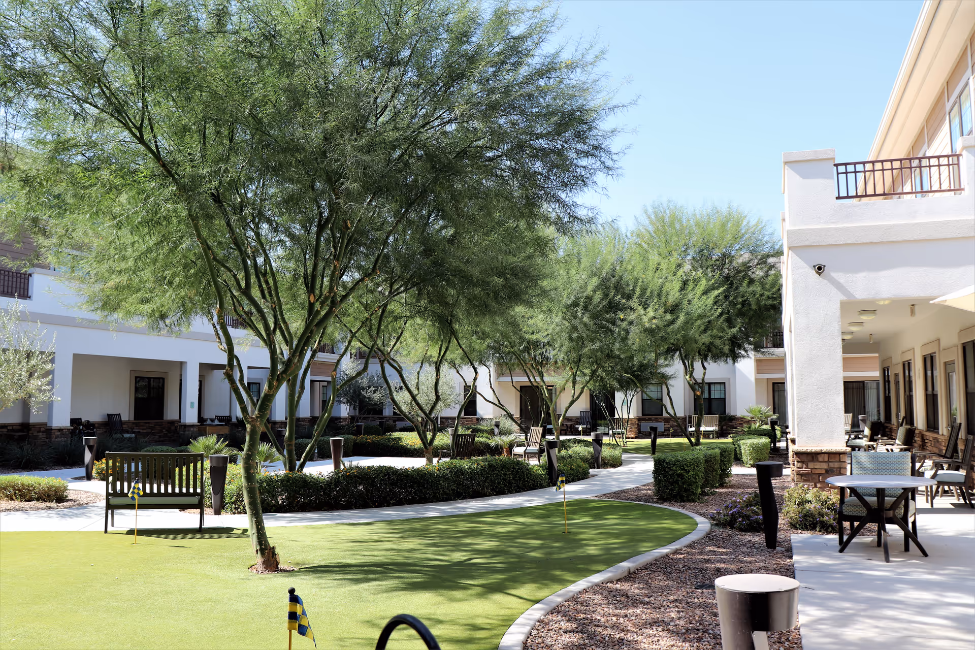 Outdoor courtyard area at Savanna House featuring a putting green with small flags, surrounded by trees, bushes, benches, and patio seating. The building has white walls with covered walkways and balconies under a clear blue sky.