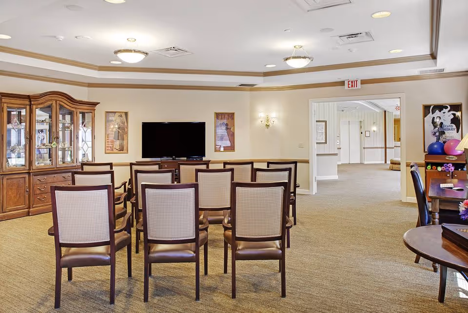 A common area in a senior living facility with rows of chairs facing a flat-screen TV mounted on a wooden cabinet. There is a large wooden display cabinet on the left side and a table with exercise balls and a lamp on the right. The room has beige walls, carpeted floor, and ceiling lights. An exit sign is visible above a doorway leading to a hallway.