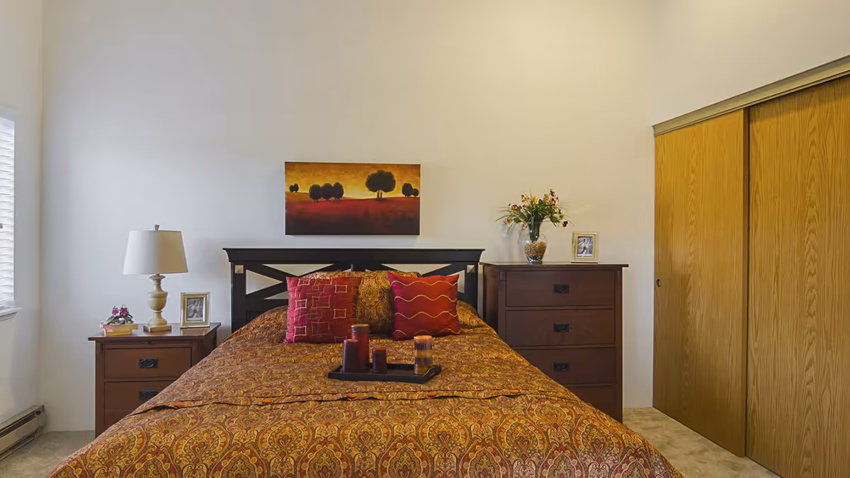 Well-lit bedroom with a patterned bedspread on a double bed, bedside tables with lamps and decor, a dresser, wall art, and sliding closet doors.
