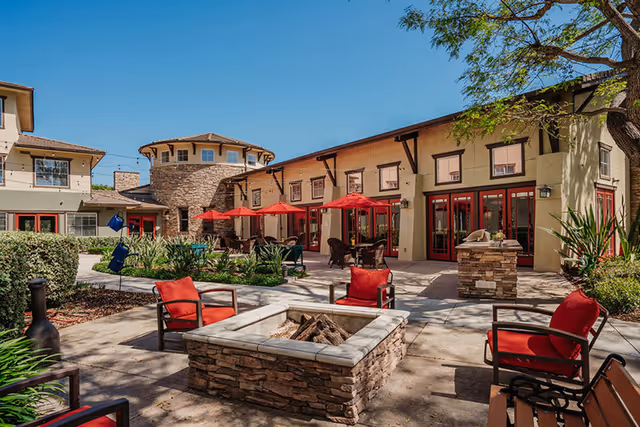 Sunny outdoor courtyard with red-cushioned chairs around a stone fire pit, tables with red umbrellas, and the surrounding two-story building.