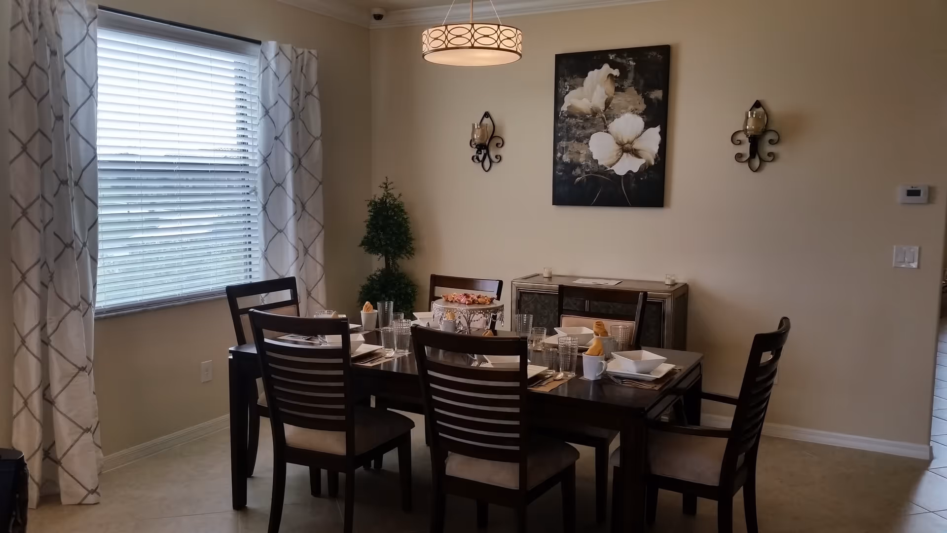 A dining room with a dark wooden table set for six people. The table has white square plates, glasses, and napkins folded in cups. There is a decorative cake stand with flowers in the center. The room has beige walls, a large window with white and gray patterned curtains, a modern ceiling light fixture, a small green plant in the corner, and a floral painting on the wall above a sideboard.