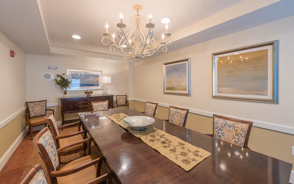 A formal dining room with a long dark wooden table surrounded by eight upholstered chairs with patterned backs. A decorative runner and a large bowl are placed on the table. The room features a chandelier hanging from the ceiling, two framed paintings on the wall, a sideboard with a lamp and a vase of flowers, and a carpeted floor.