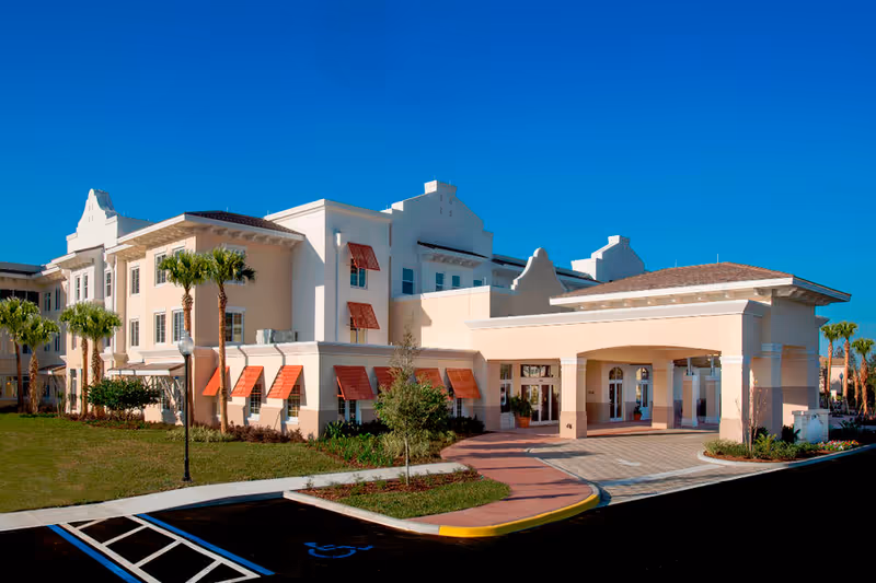 Exterior view of Encore at Avalon Park Assisted Living and Memory Care Community, showing a large, light-colored building with red awnings over the windows, palm trees, a covered entrance, and a clear blue sky.