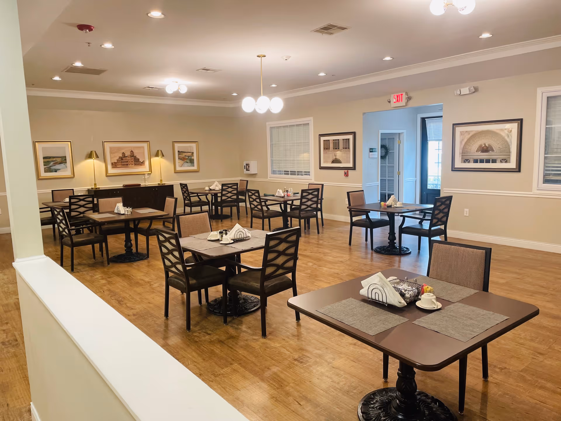 A clean and well-lit dining room with several square tables, each set with placemats, napkins, and condiments. The room has wooden flooring, beige walls adorned with framed artwork, and ceiling lights. There are multiple chairs around each table, and an exit door is visible in the background.