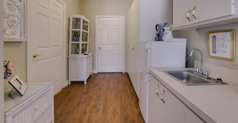 Hallway-style kitchenette with a sink and cabinets on the right, wood flooring, and storage furniture and decorative items along the left.