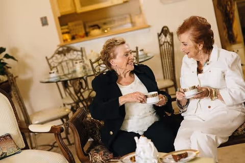 Two elderly women sitting in a cozy living room area, smiling and holding teacups while engaging in conversation. The room features a glass dining table with chairs in the background and warm, inviting decor.